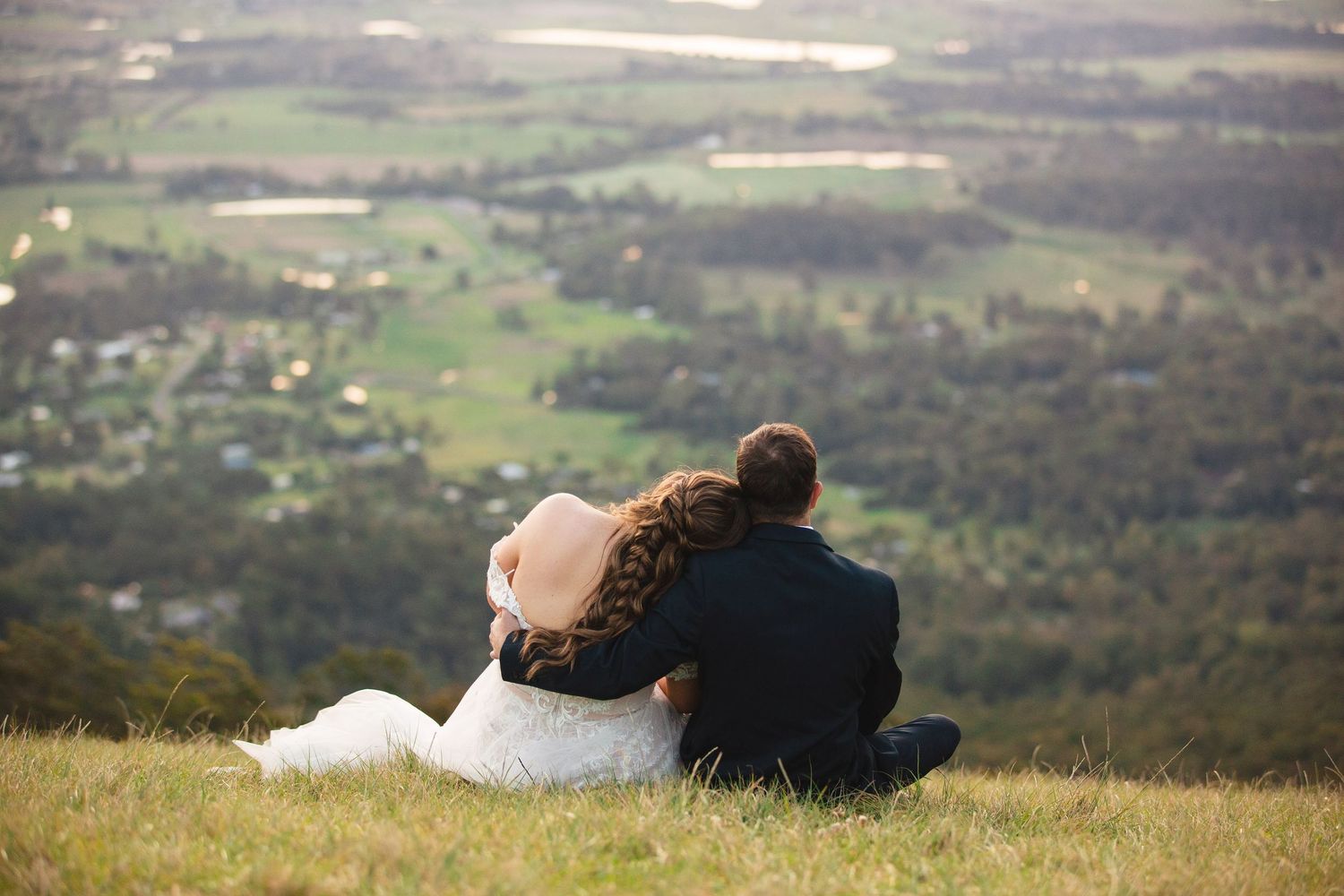A couple sits close together on a grassy hilltop overlooking a scenic valley landscape on a peaceful afternoon.