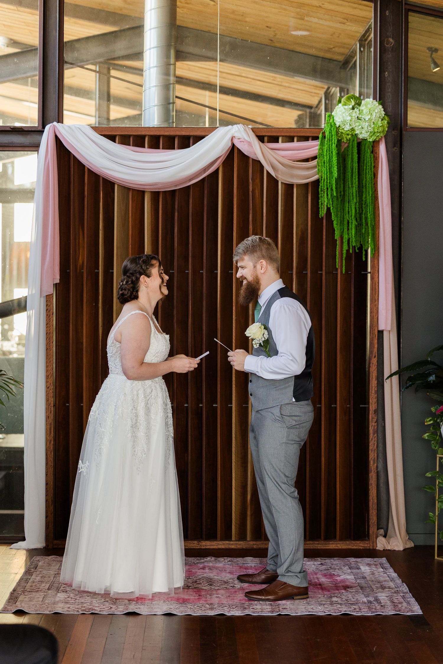 Bride and groom exchange vows indoors at decorated wooden backdrop with pink draping and greenery.