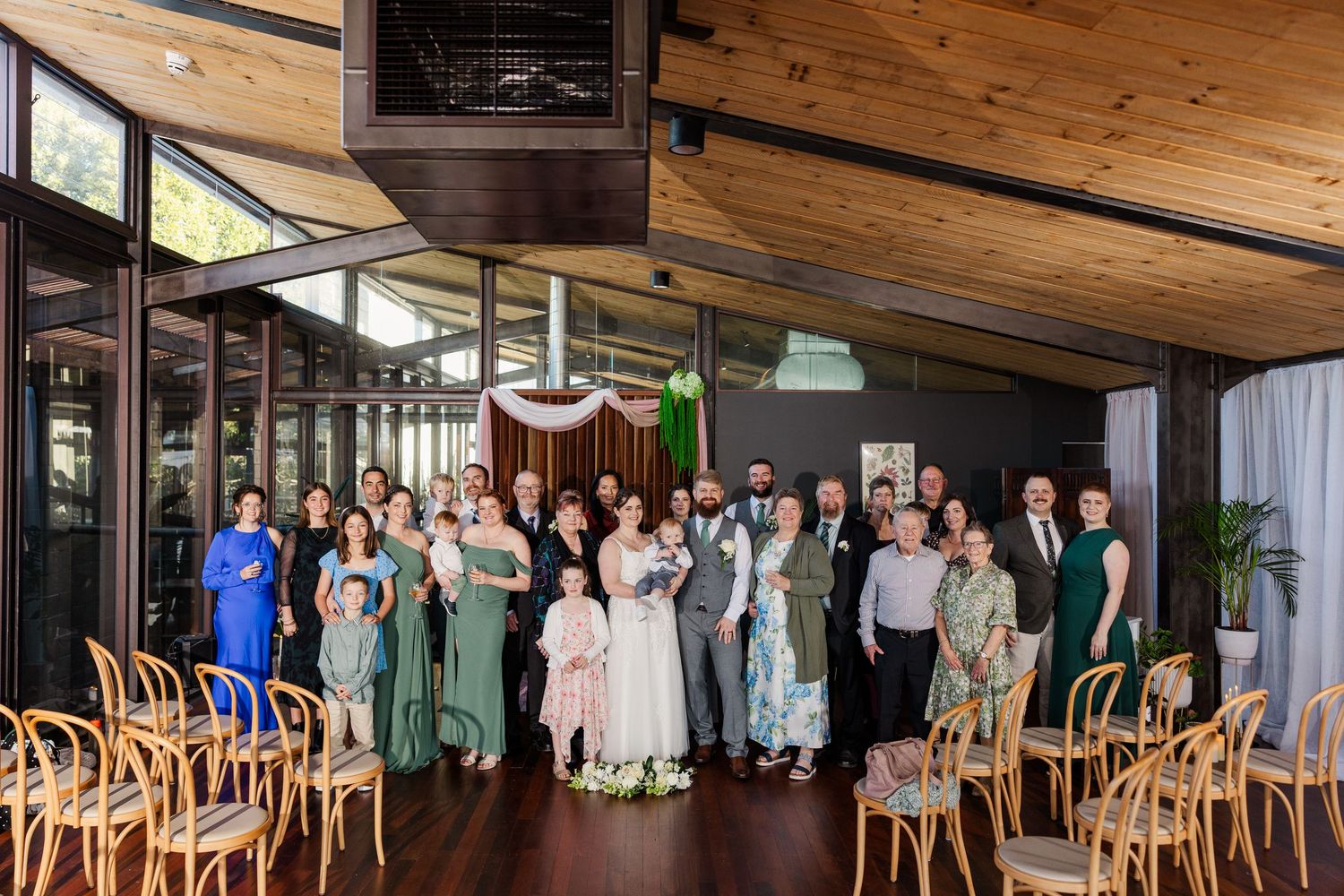 Wedding party and guests pose together in a modern venue with wooden ceiling and large windows.