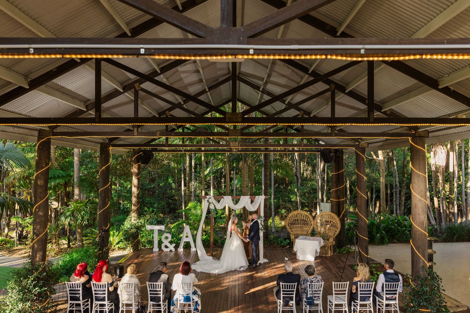 Outdoor wedding ceremony under a rustic pavilion surrounded by lush greenery with guests seated in white chairs.
