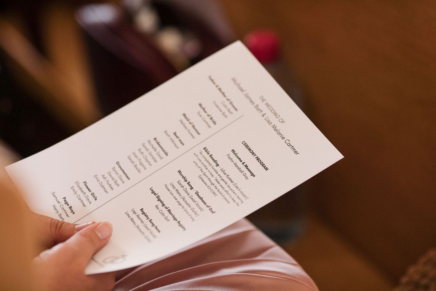 Person holding a white folded wedding program with printed text at an indoor ceremony.