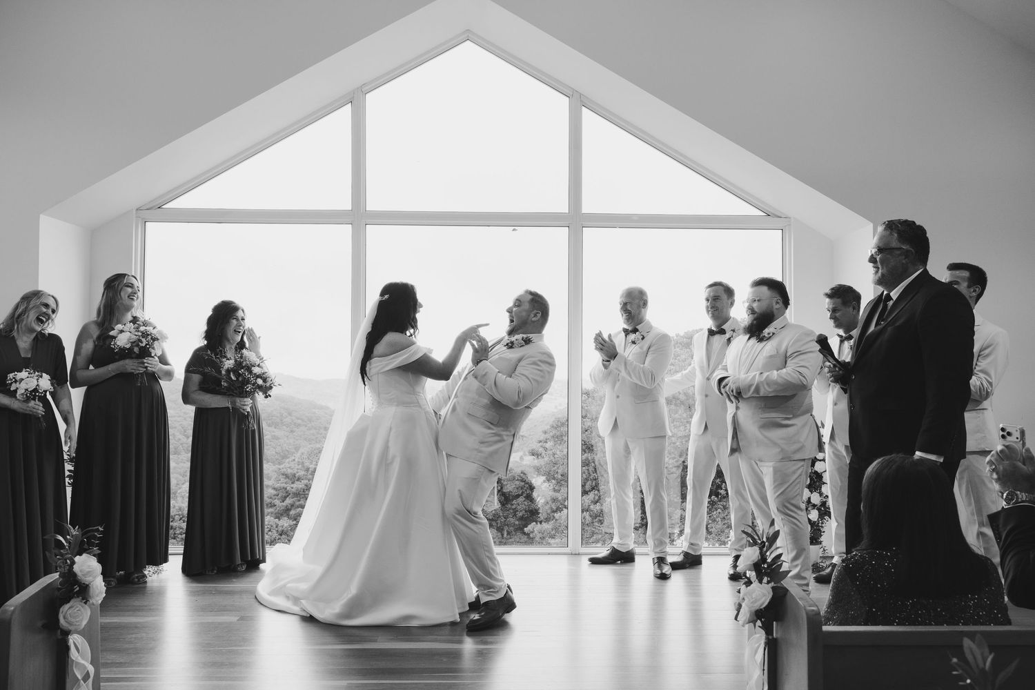 Black and white photo of bride and groom sharing first dance in chapel with large window backdrop and wedding party.