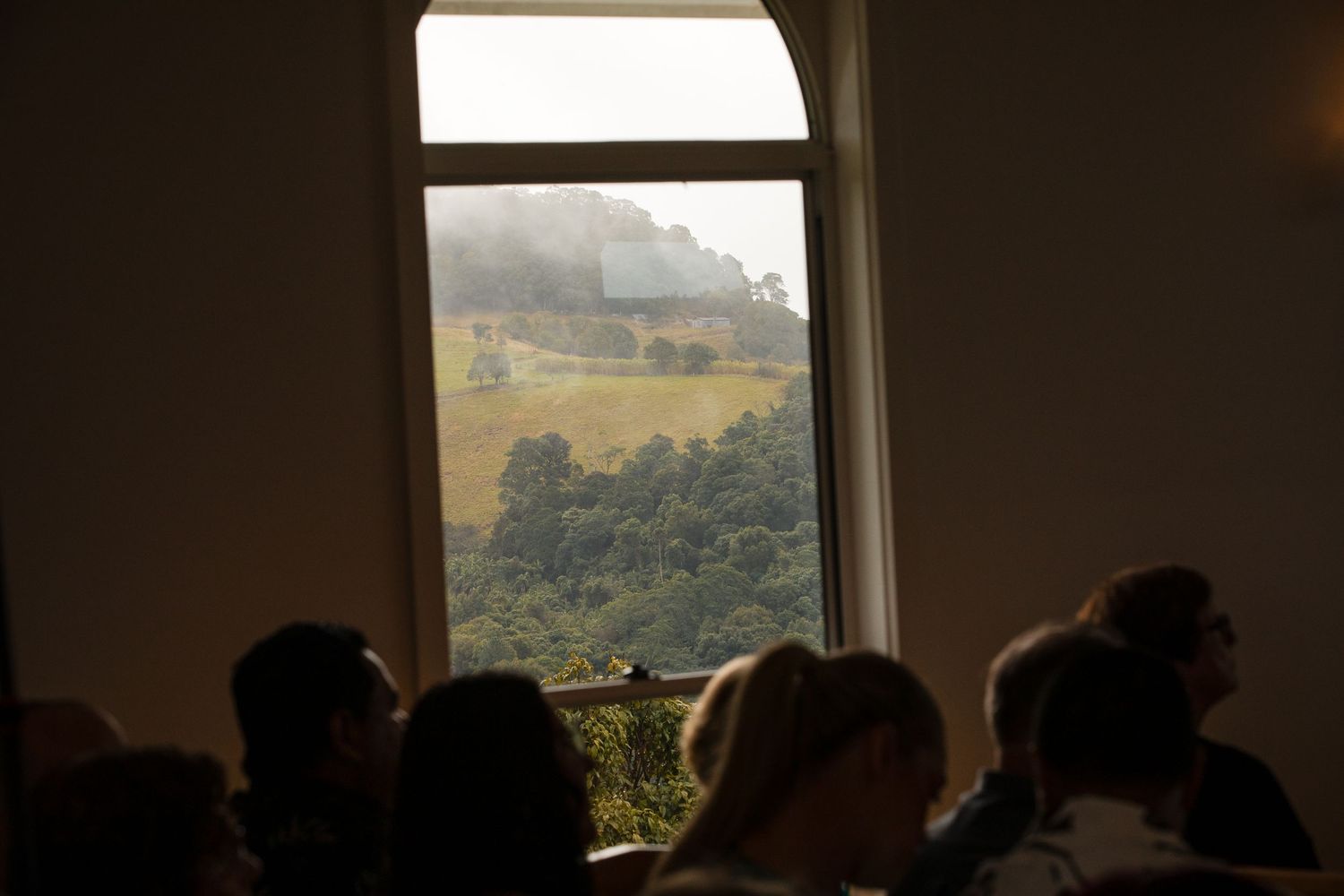 People seated indoors viewing a scenic landscape through a large window with misty hills and trees.