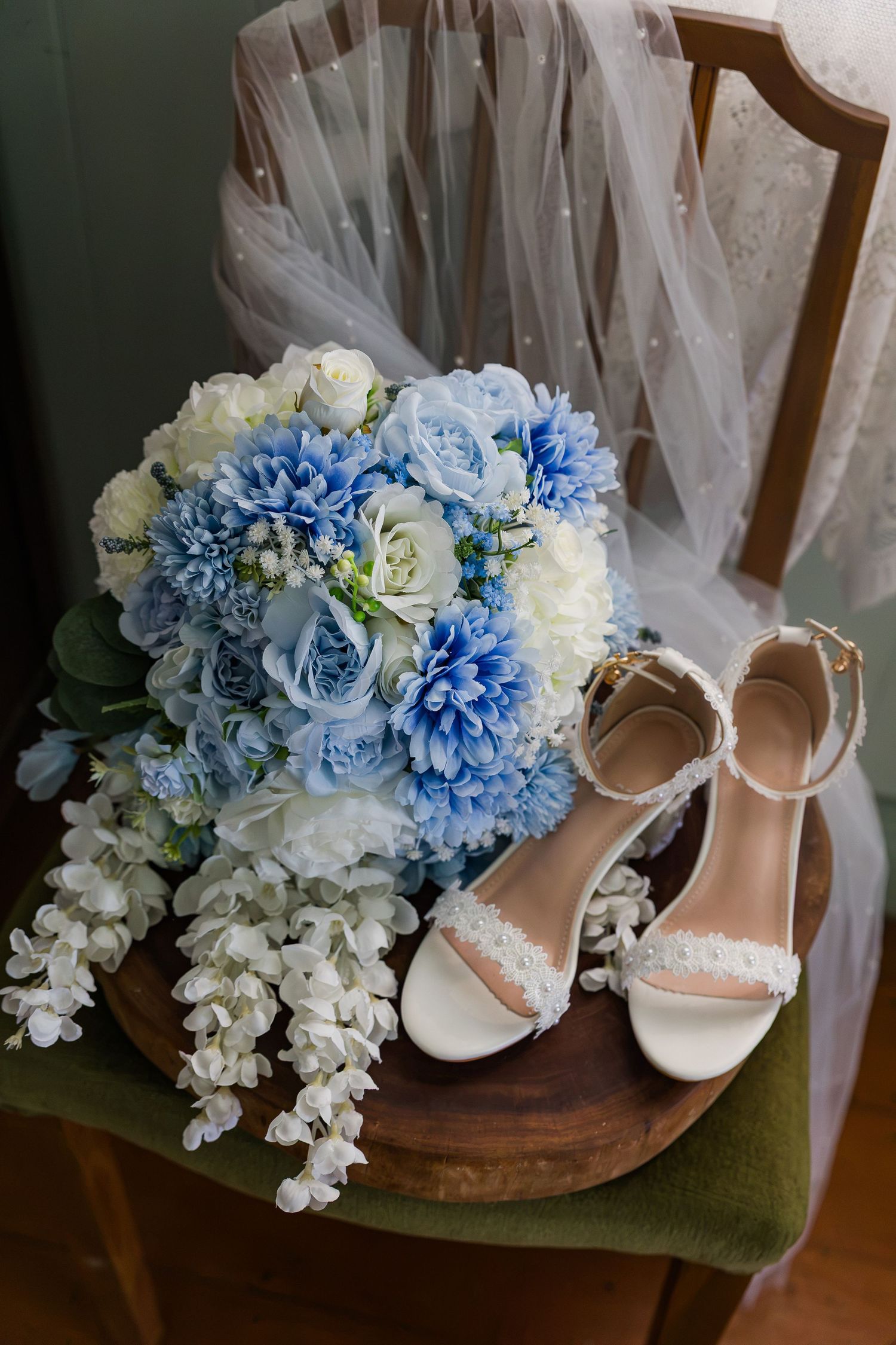 White bridal heels with ankle straps beside a blue and white wedding bouquet on a wooden chair with veil.