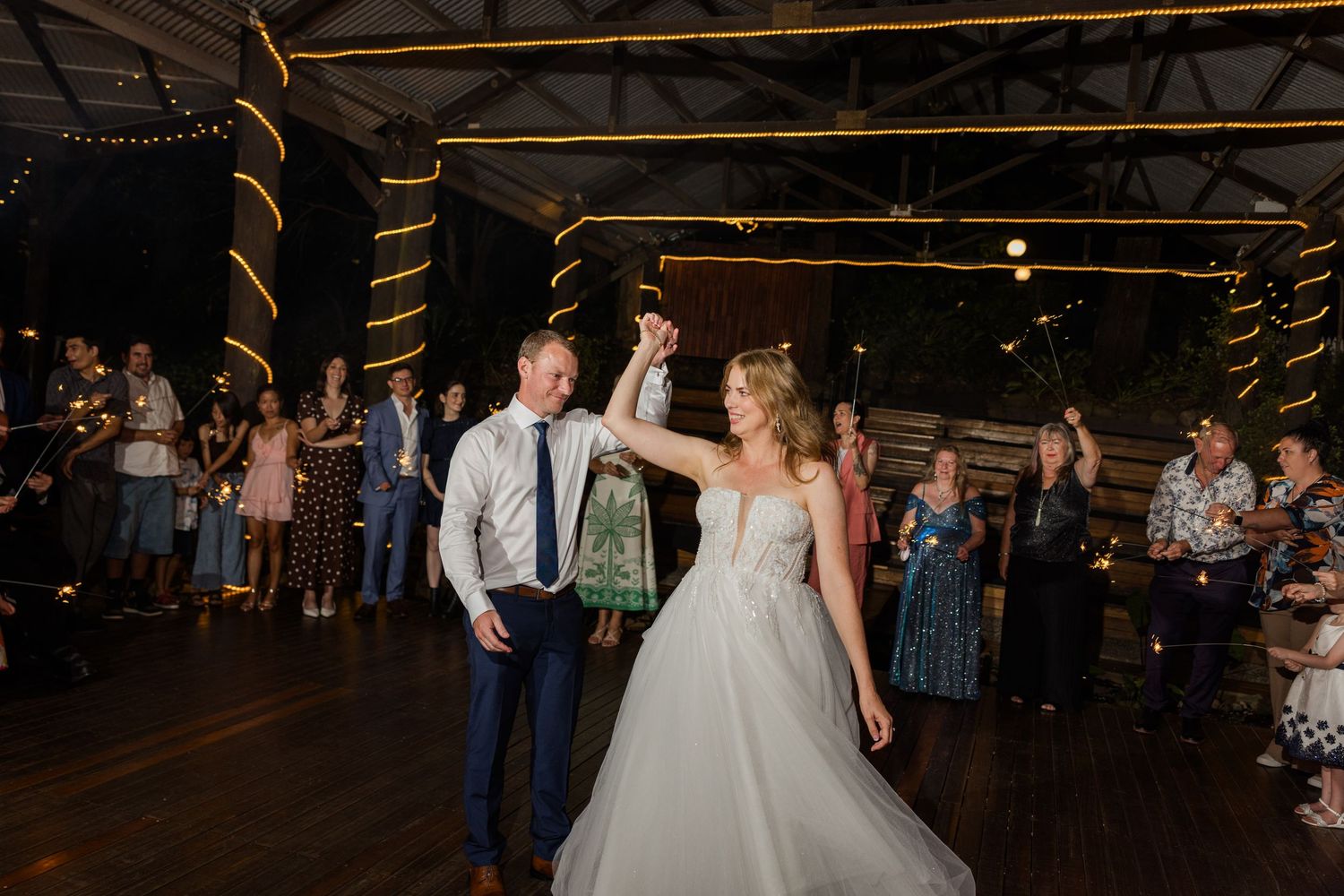 Wedding guests dance together on a rustic wooden dance floor under warm lighting at an evening reception.