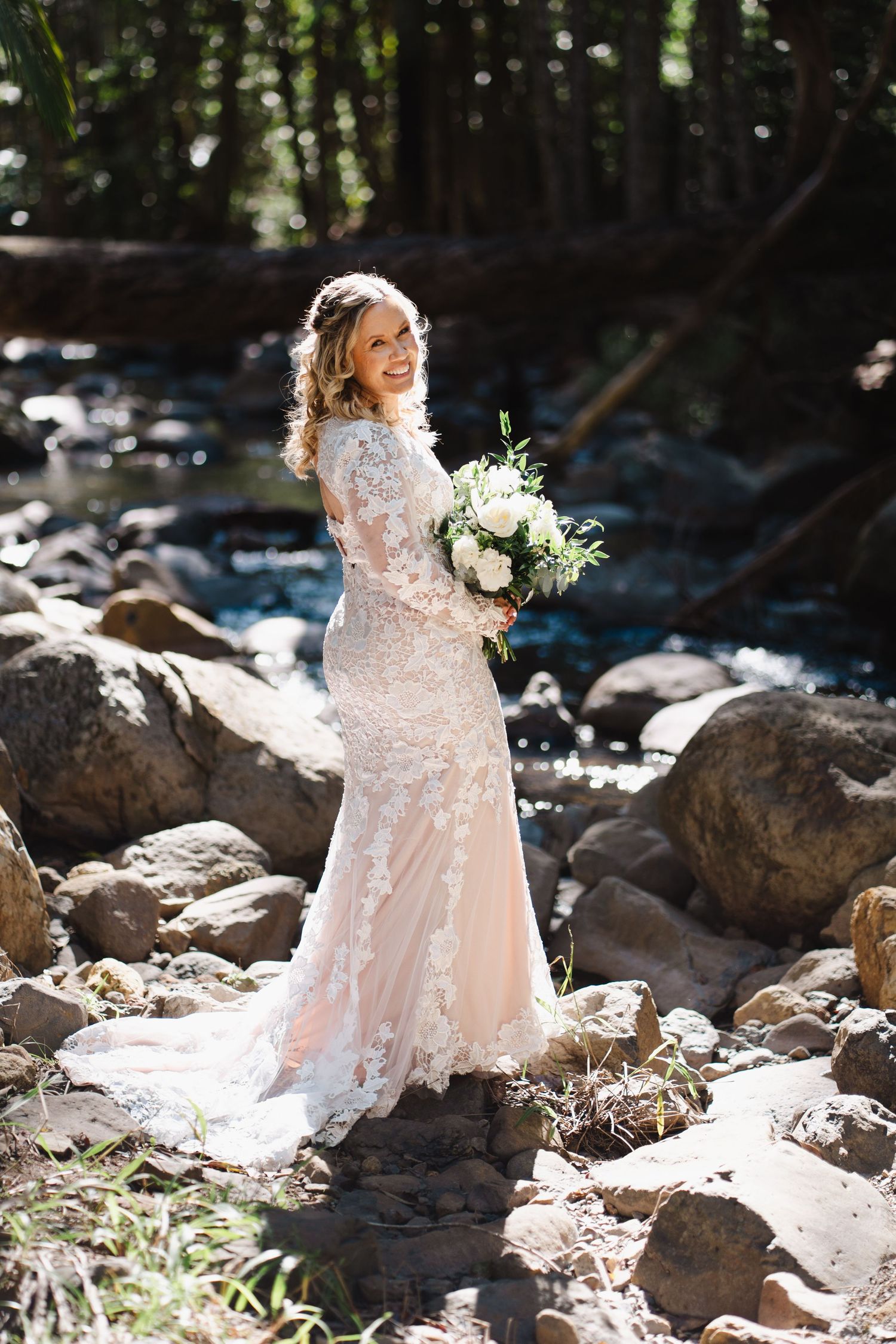 A bride in a lace wedding dress holds a white bouquet while standing on rocky terrain near a stream in dappled sunlight.