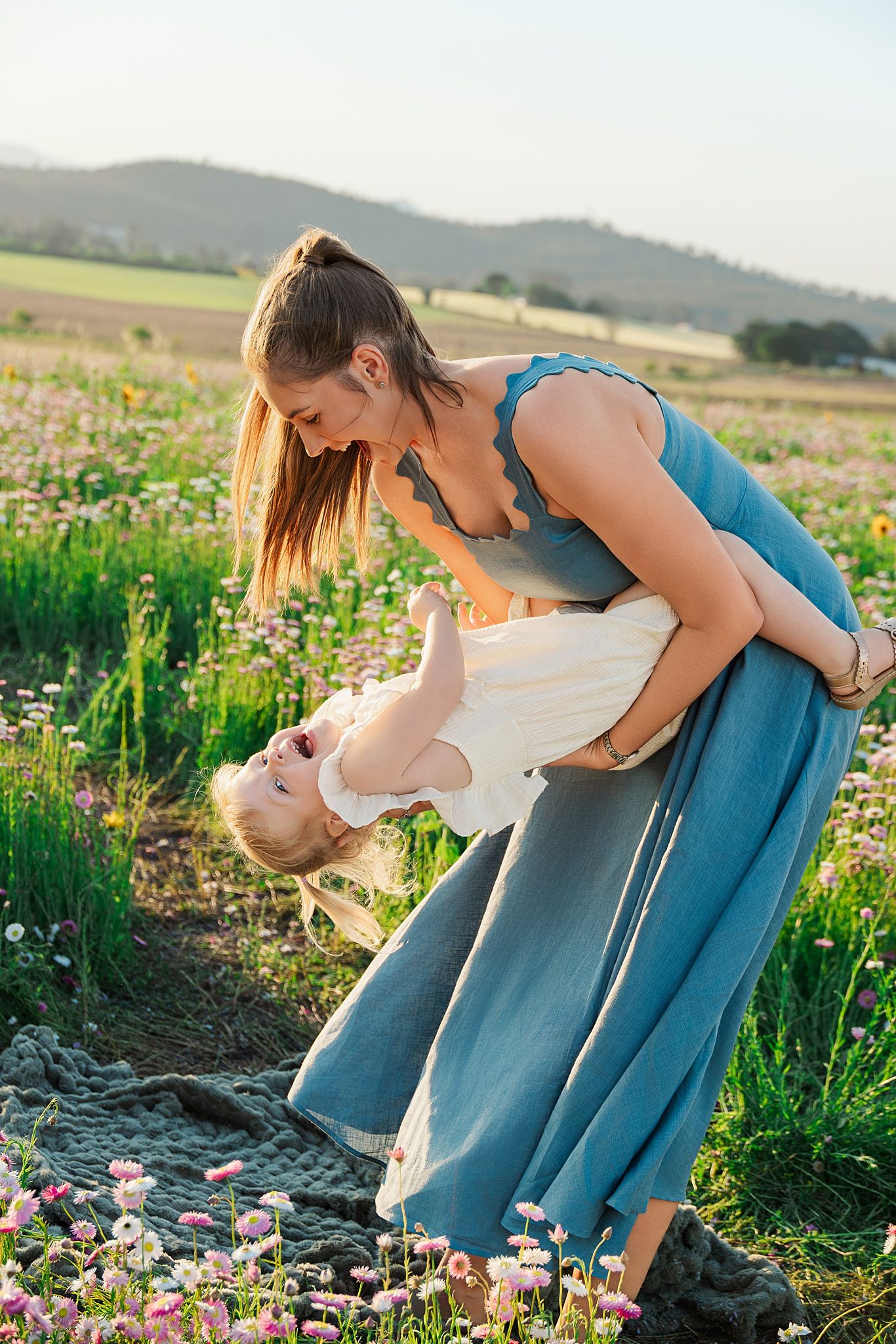Smiling woman in a blue dress playfully tips a laughing toddler sideways in a sunny wildflower field with rolling hills behind.