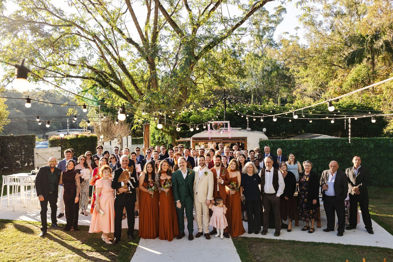 Large wedding group photo outdoors under trees with string lights, guests in formal attire on green lawn.