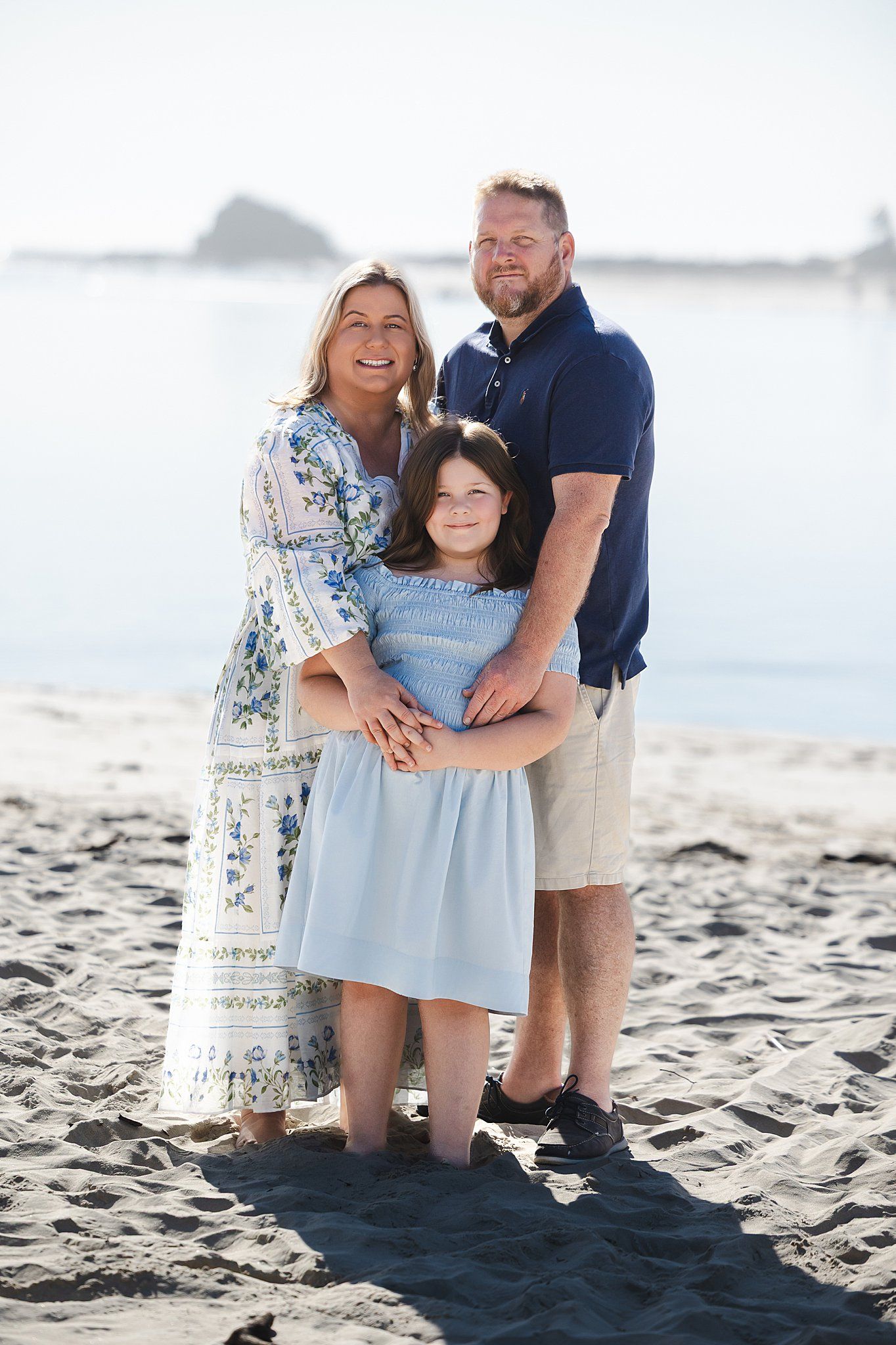 A family poses together for a portrait on a sunny beach with ocean views in the background.