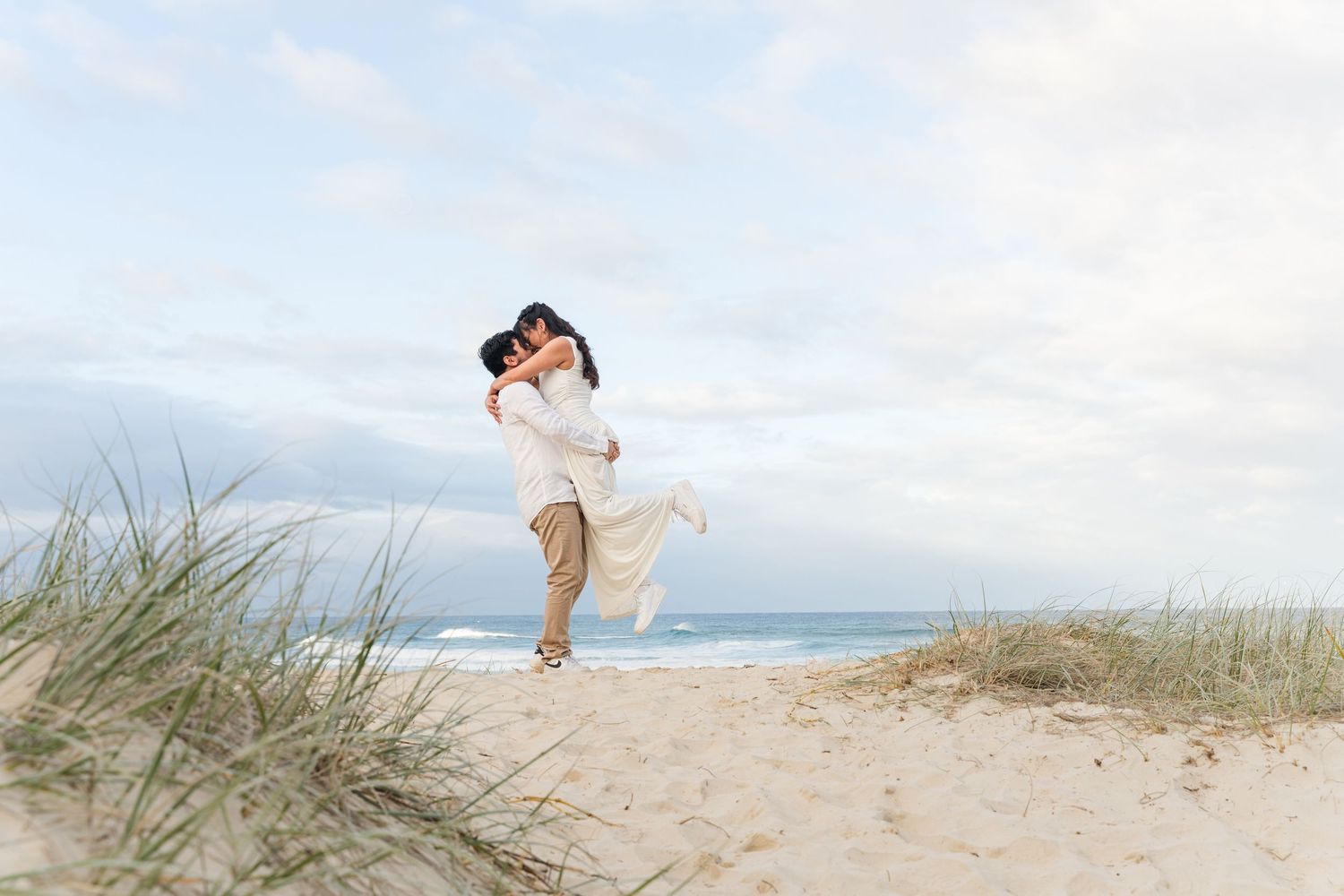 Couple embracing joyfully on a sandy beach with ocean waves and grass dunes under a cloudy sky.