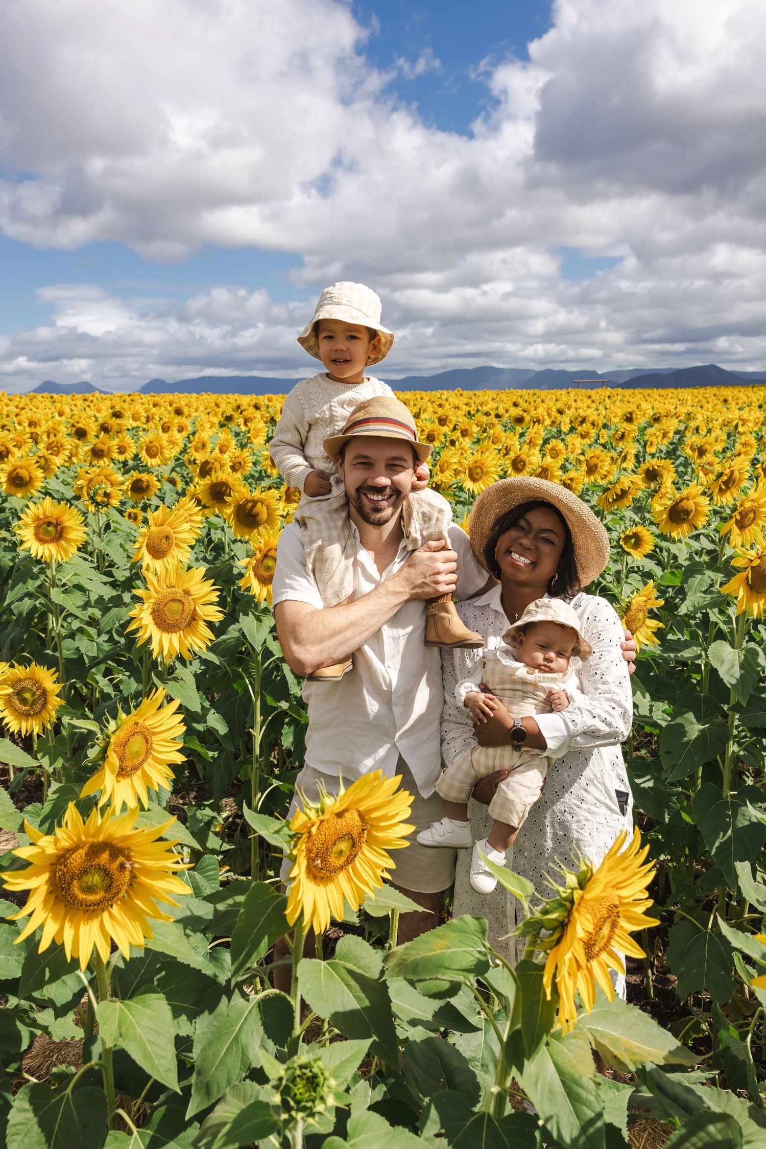 Happy interracial family with two young children posing in a vast sunflower field under a partly cloudy sky.