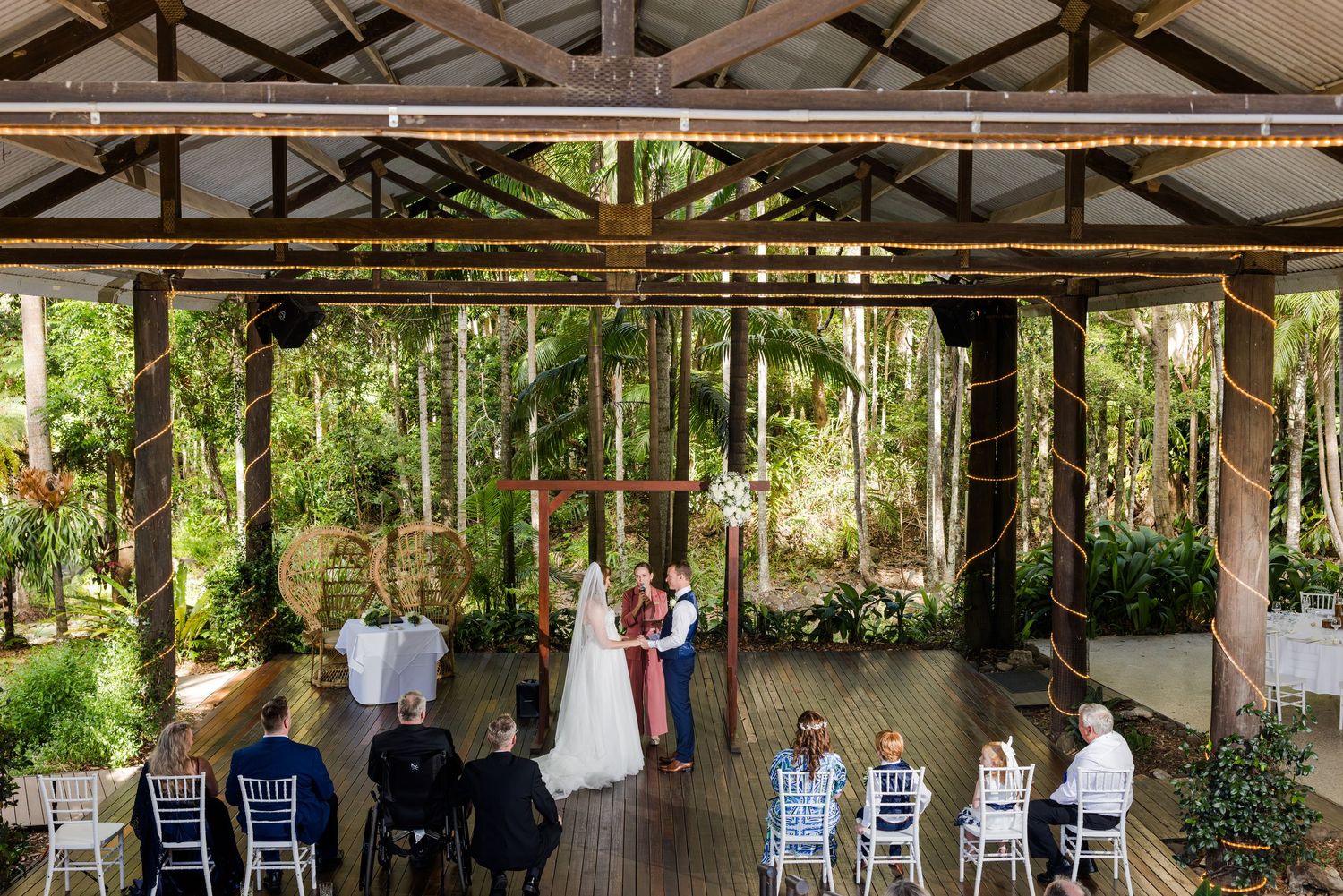 Intimate outdoor wedding ceremony under a rustic wooden pavilion surrounded by lush tropical gardens and palm trees.
