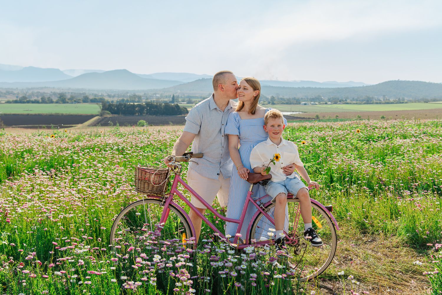 Family with child posing by pink bicycle in blooming flower field with mountain views on sunny day.