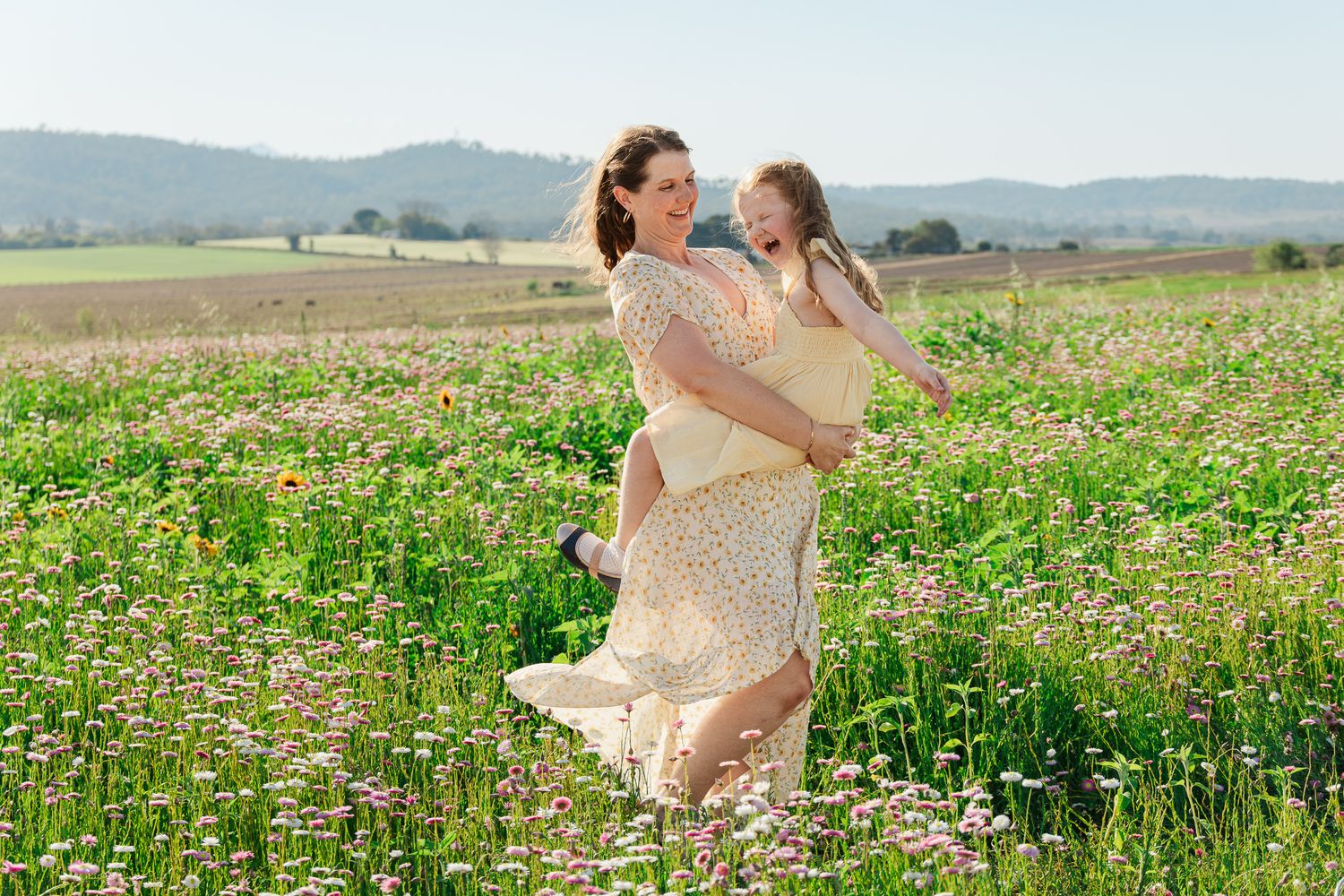 Mother holding daughter in a sunny wildflower meadow, both wearing yellow dresses with rolling hills behind.