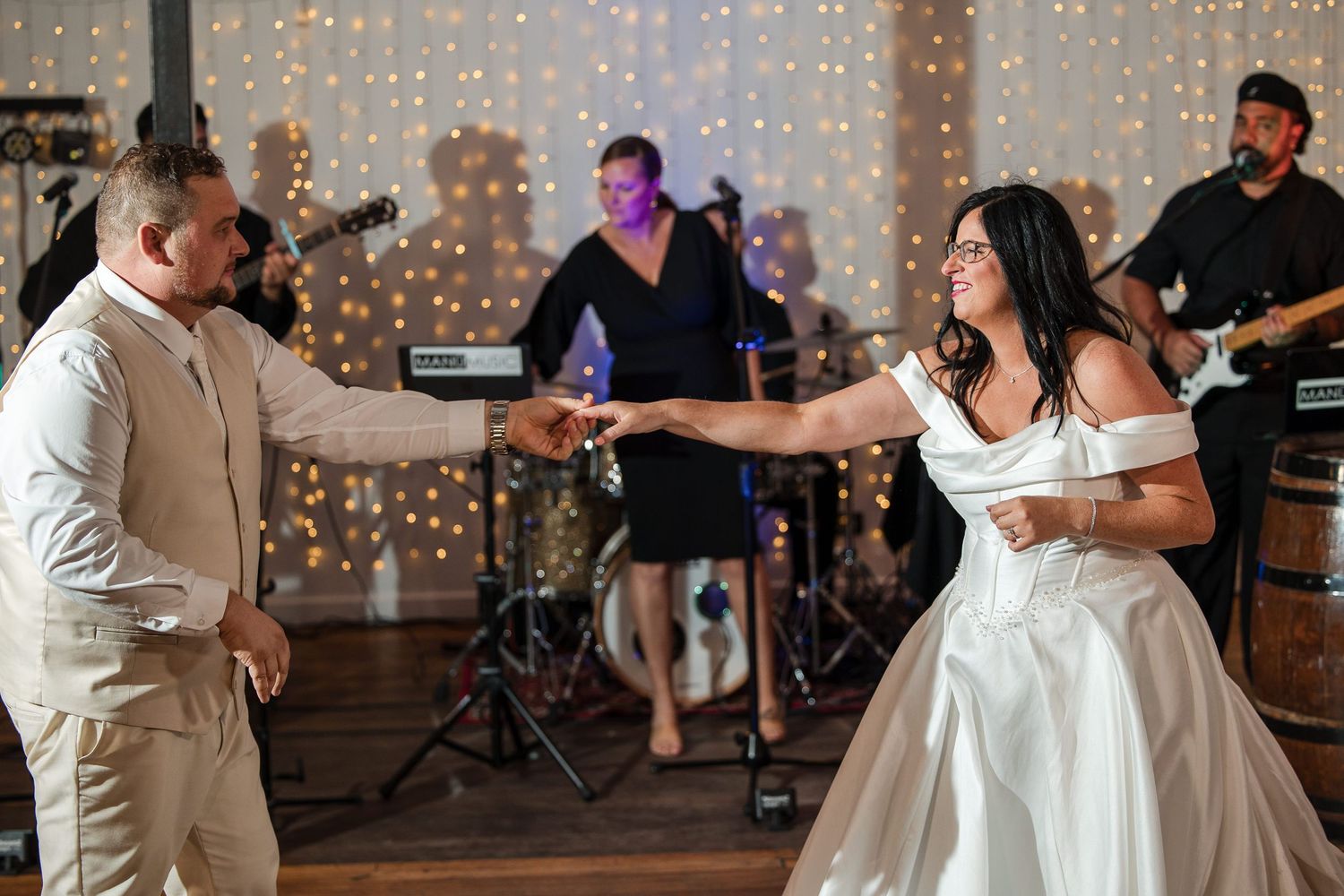 Bride and groom share first dance at wedding reception with live band performing and fairy lights in background.
