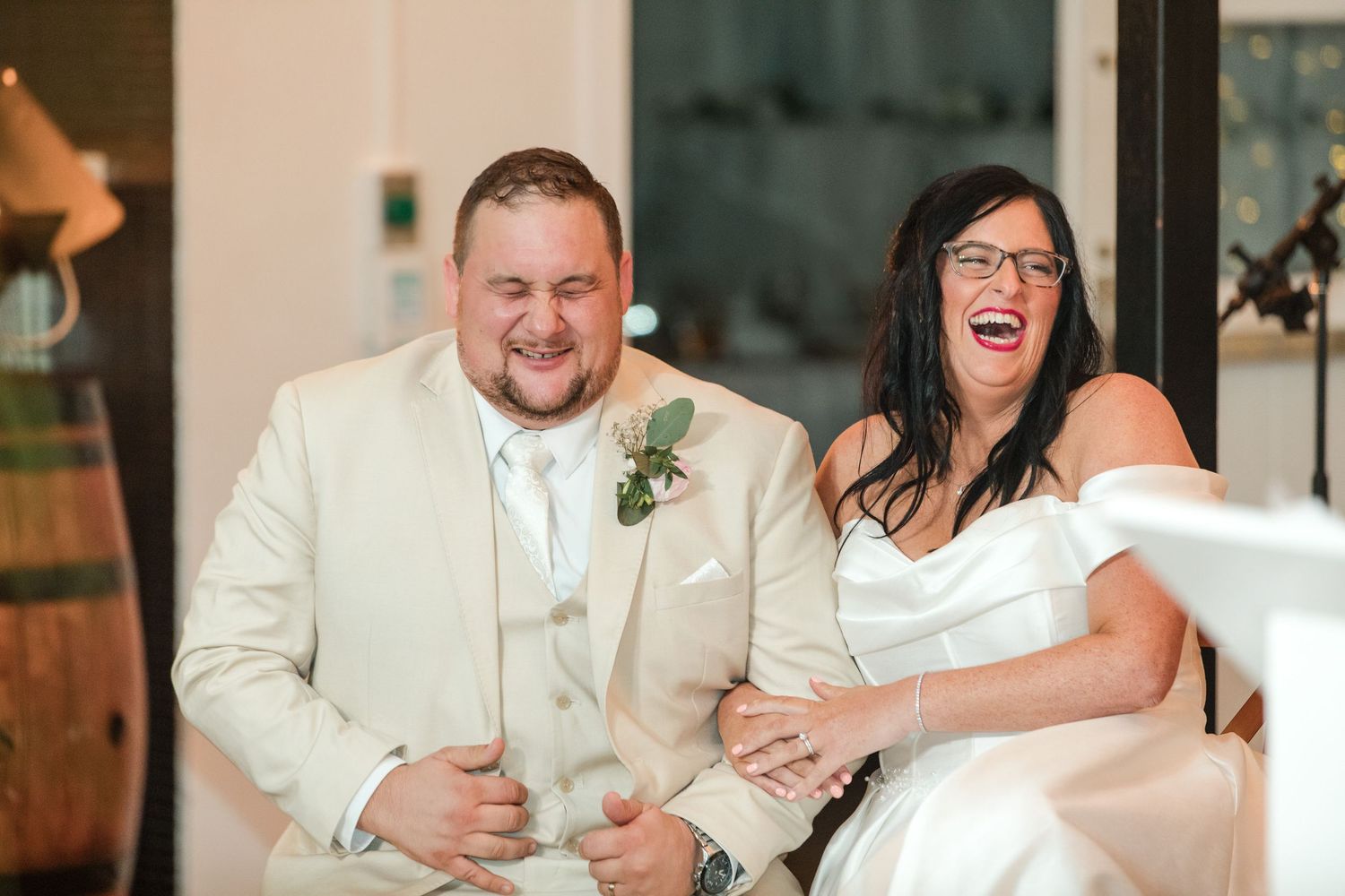 Bride and groom laughing joyfully during wedding reception, groom in beige suit, bride in white off-shoulder dress.