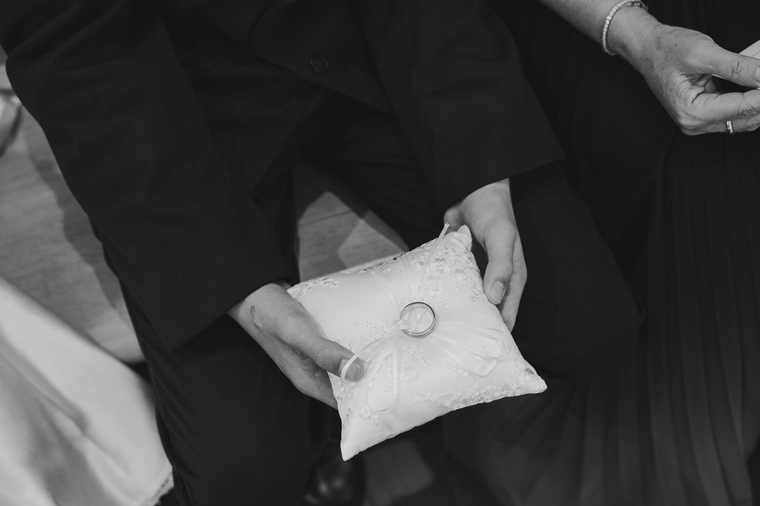 Black and white photo of wedding rings on a white pillow held by hands during a wedding ceremony.