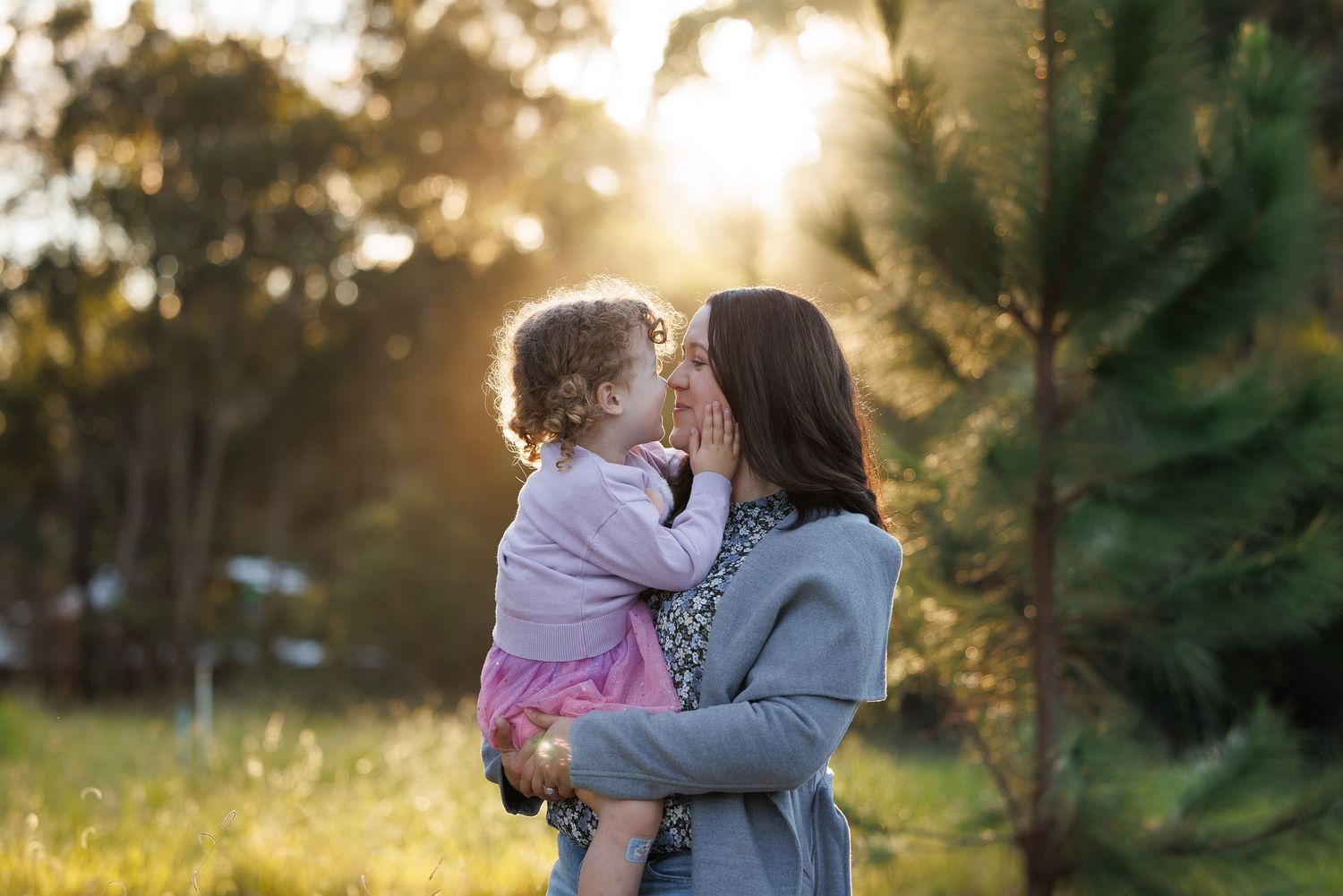 A mother holds her toddler daughter, sharing a tender kiss outdoors at golden hour near pine trees.