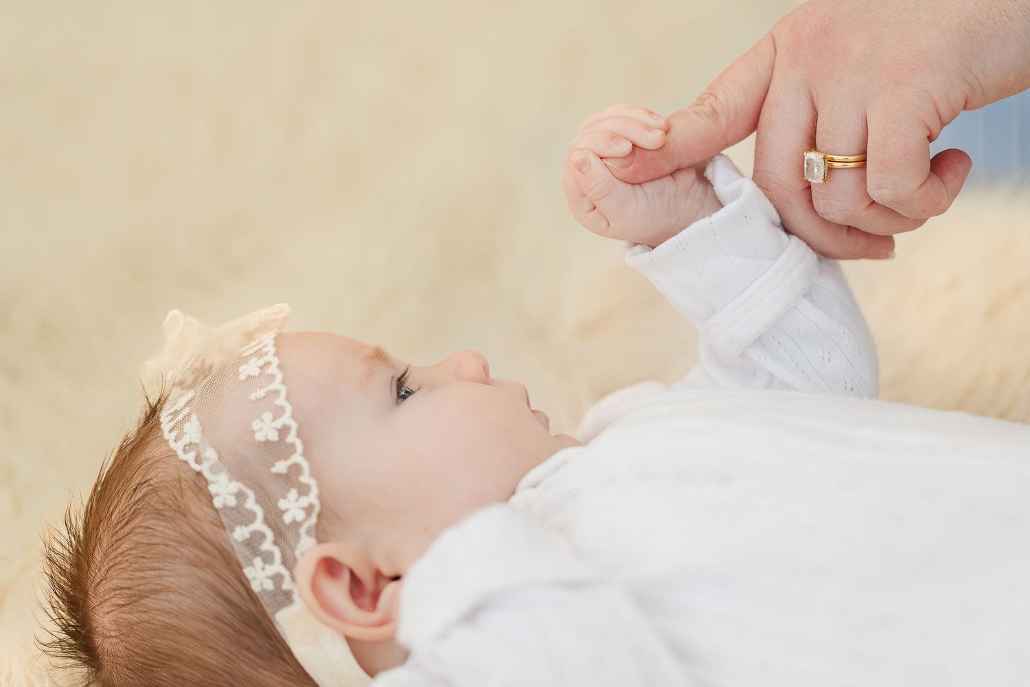 A baby in a white outfit and lace headband gently holds an adult's finger while lying on a soft surface.