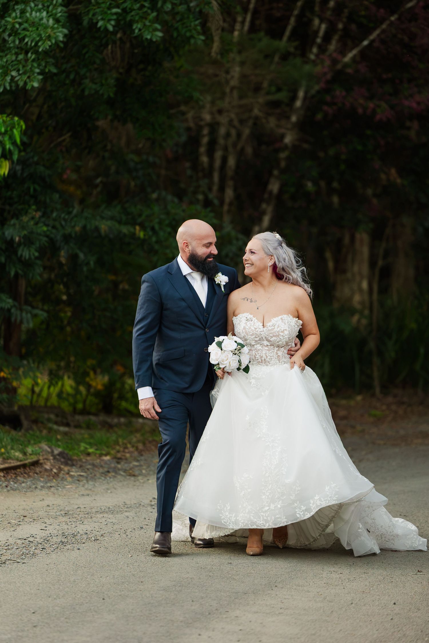 Bride and groom walking together outdoors, she in a lace ballgown, he in a navy suit, smiling at each other.