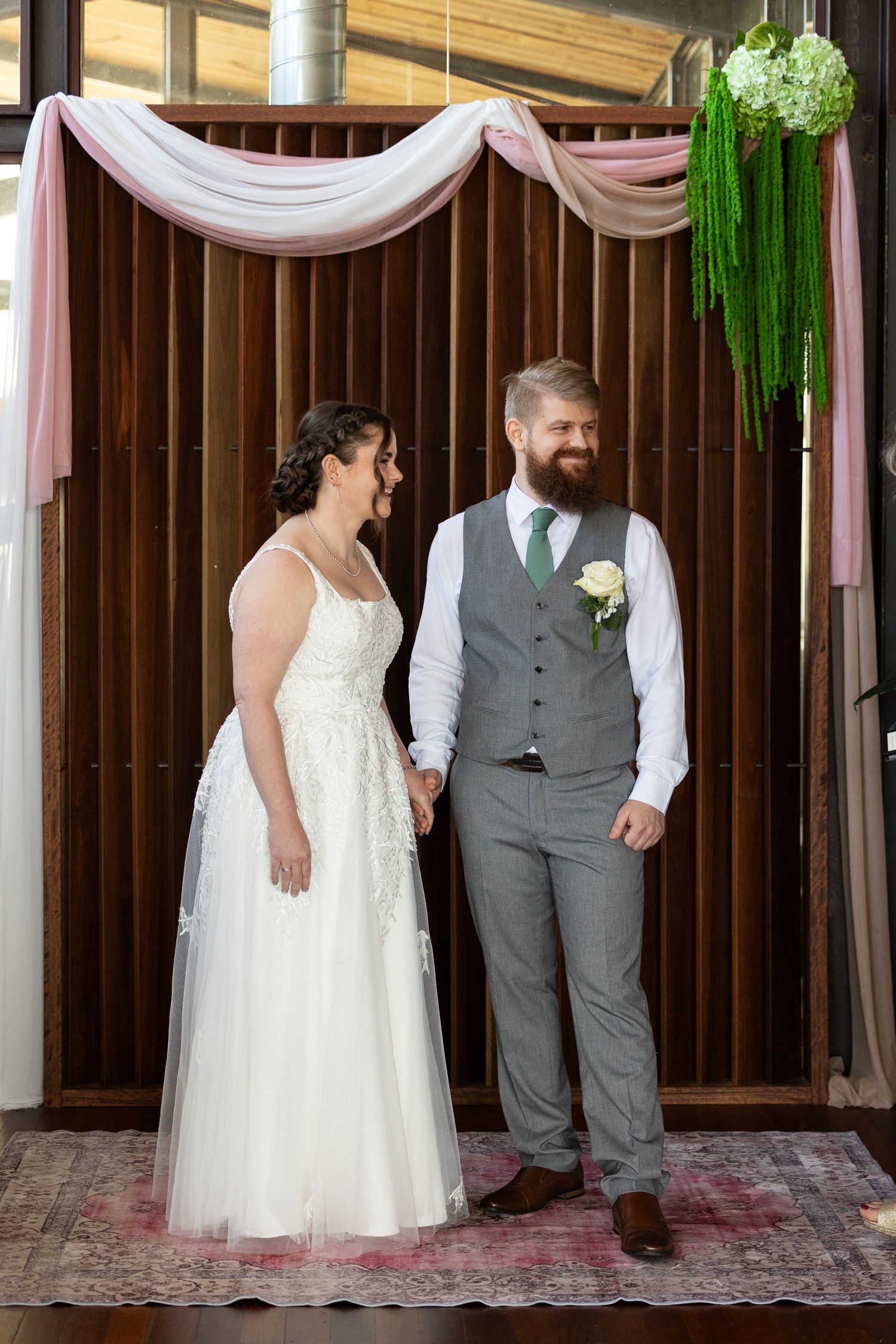 Bride in white gown and groom in gray vest hold hands smiling at each other under floral arch in rustic venue.
