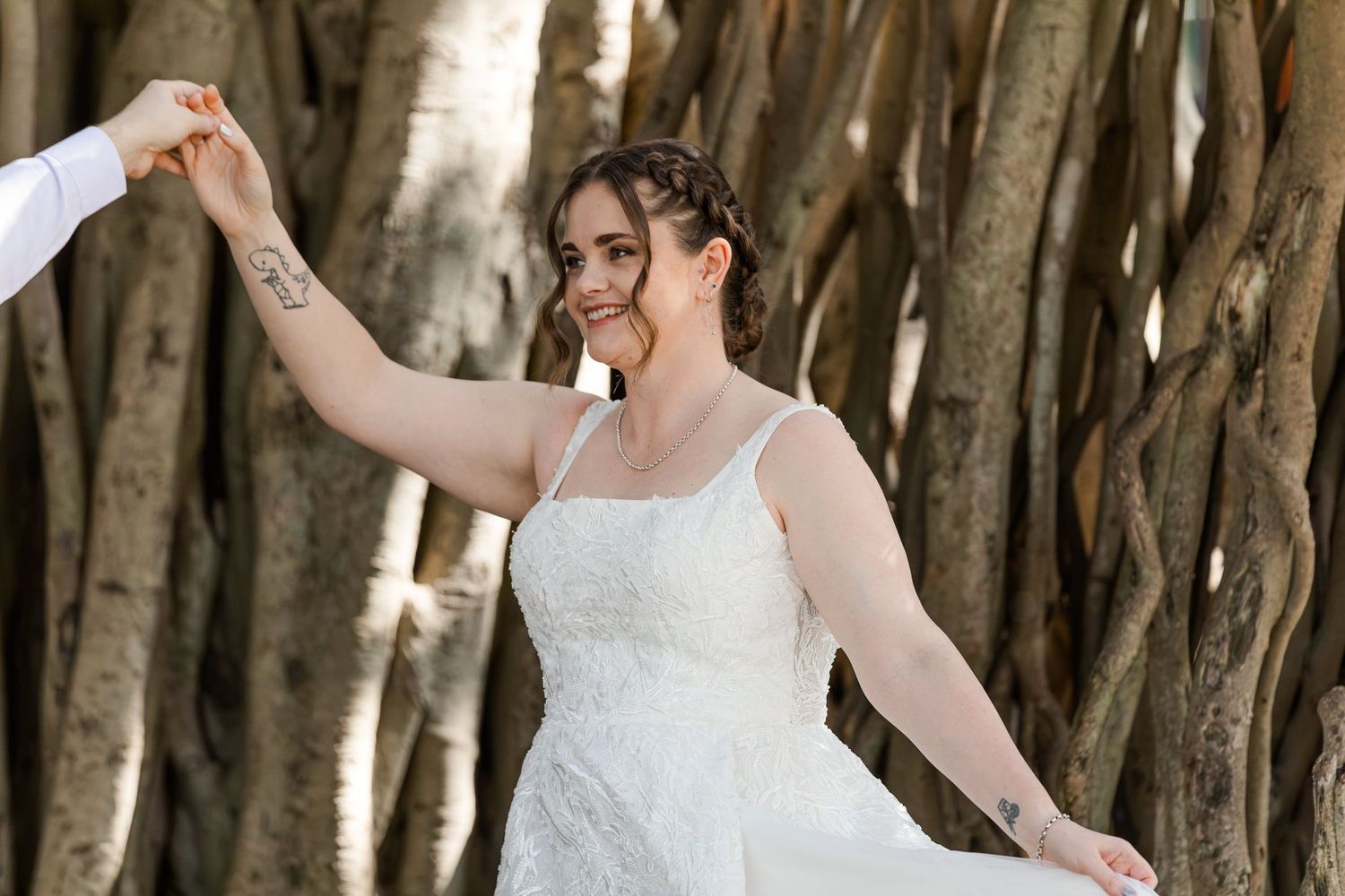 Bride in white lace dress dancing outdoors near banyan tree roots, smiling joyfully while holding hands.