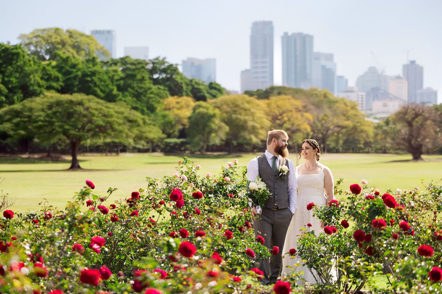 Bride and groom stand among red roses in a lush park with city skyline backdrop on their wedding day.