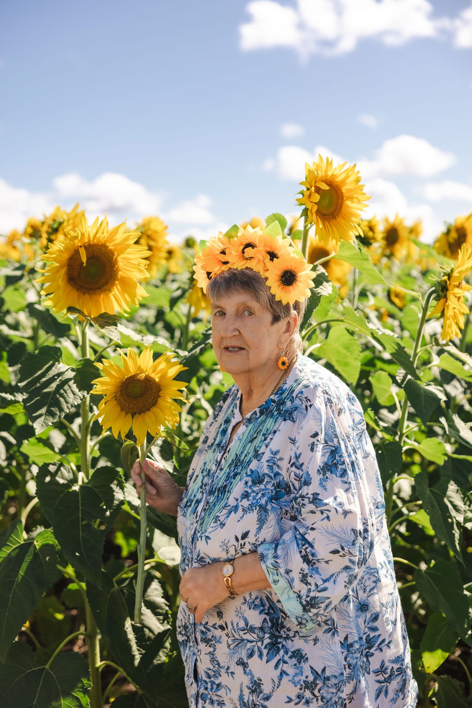 Elderly woman wearing sunflower crown holding sunflower in a vibrant sunflower field under blue sky.