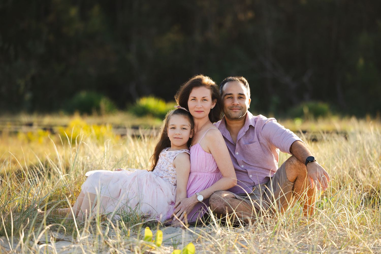 Family of three sitting together in a golden grass field, smiling warmly in soft natural outdoor lighting.
