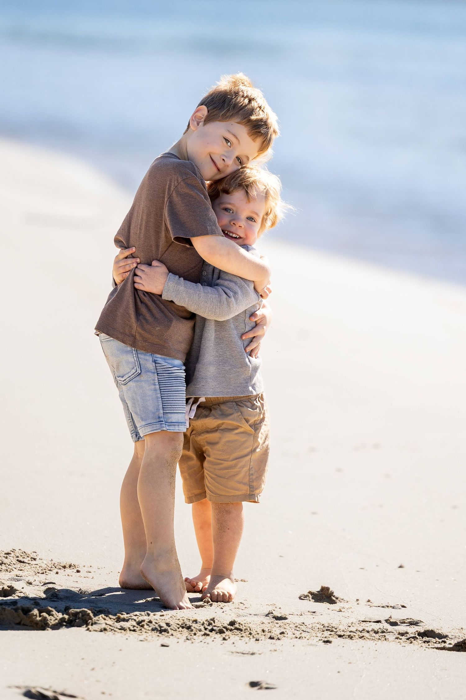 Two young brothers hugging and smiling on a sandy beach with ocean waves in the background.