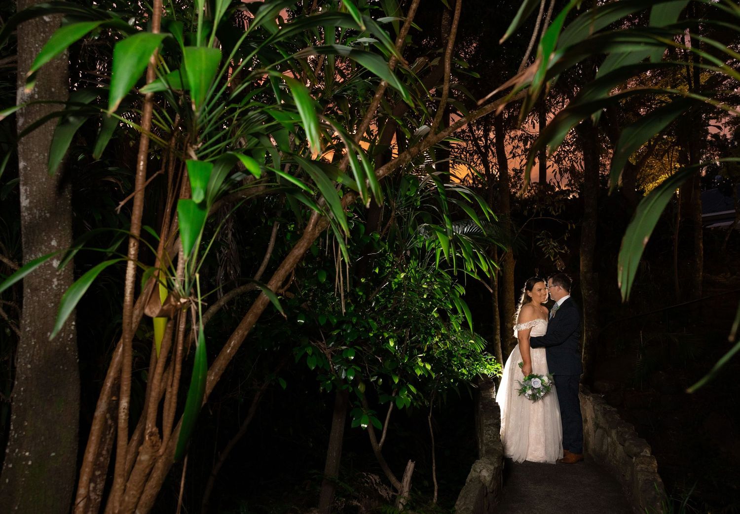A couple stands together in a romantic garden setting at night illuminated by warm lighting among tropical foliage.