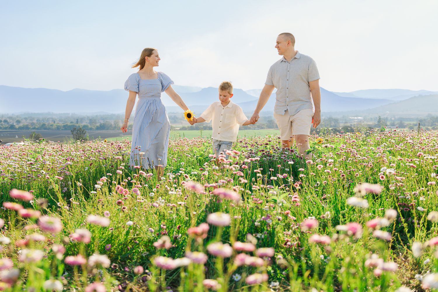 Family of three walking through a blooming wildflower meadow with mountains in the background on a sunny day.