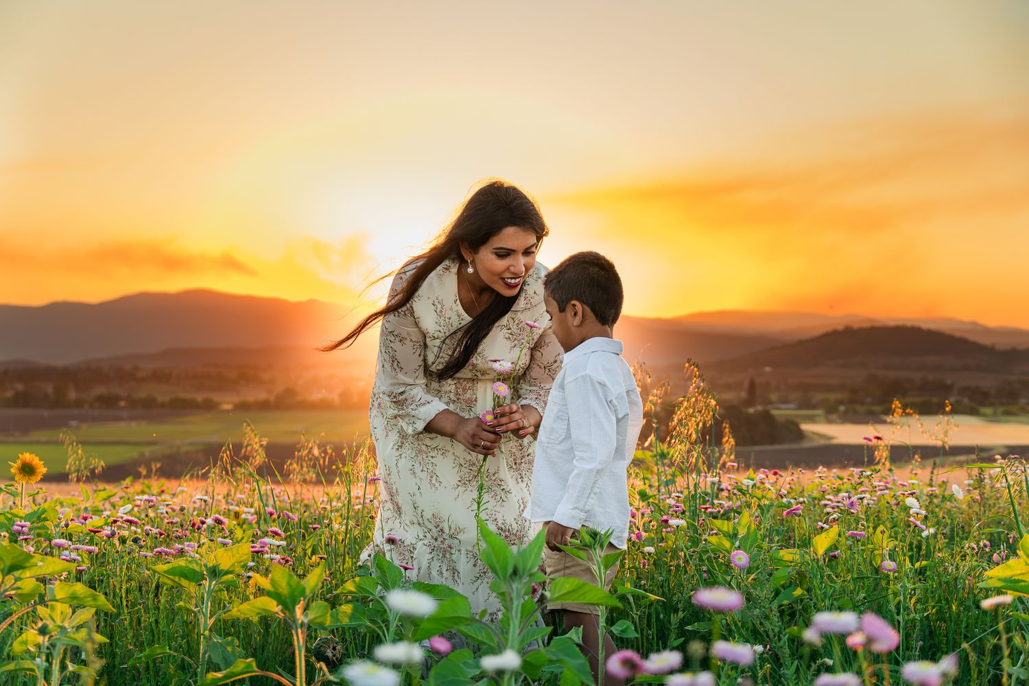 Mother and son share a tender moment in a blooming flower field at golden sunset with mountain views.