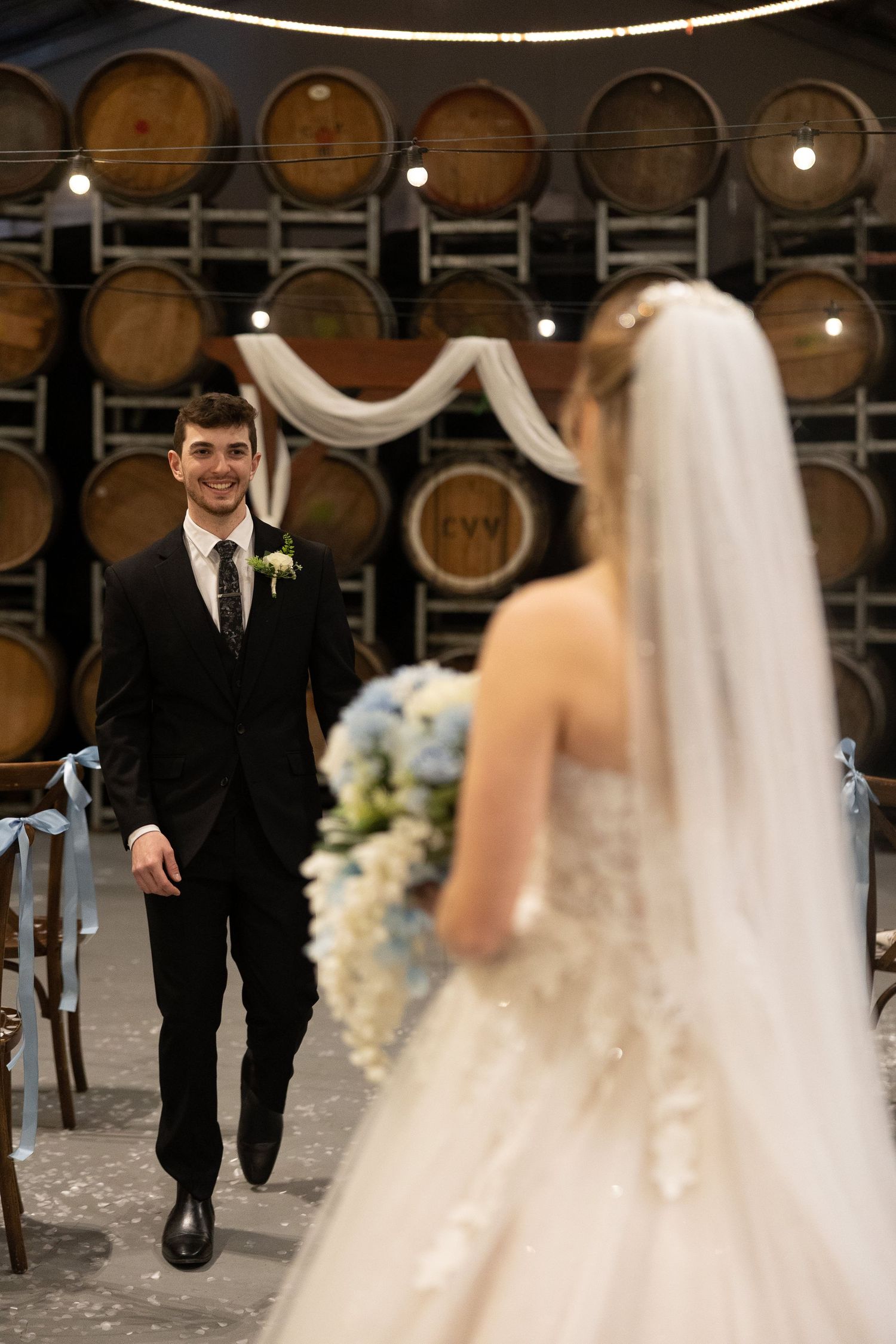Groom in black suit smiles at bride in white gown during wedding ceremony in rustic winery with barrel backdrop.