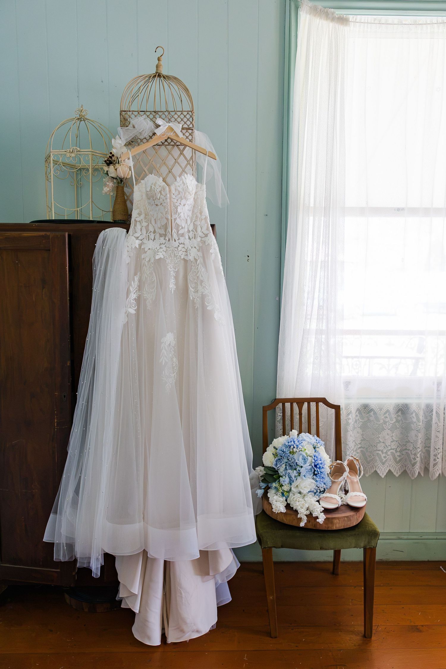 Elegant lace wedding dress hanging on a birdcage stand with blue hydrangea bouquet and bridal heels on a vintage chair.