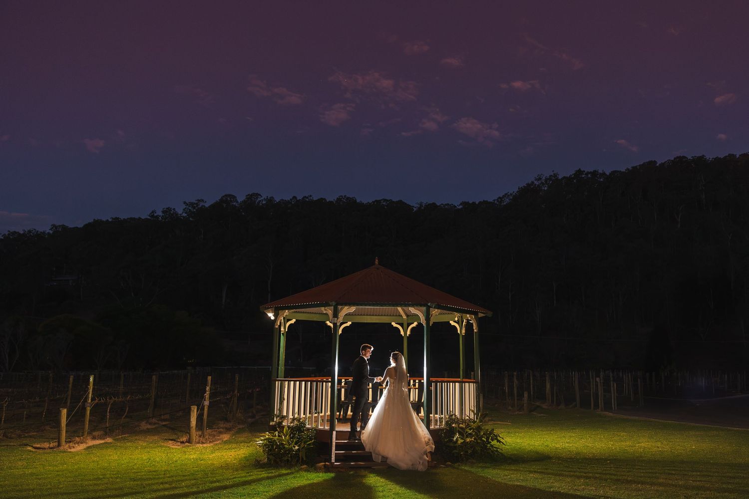 Bride and groom standing in a lit gazebo at dusk with a purple sky and lush green surroundings at an outdoor wedding.