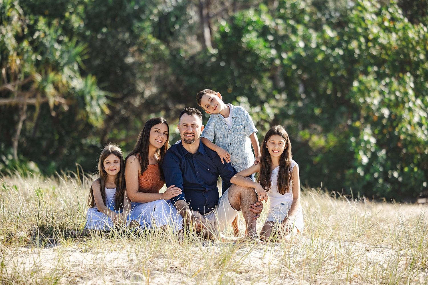 A happy family sits together in tall grass on a sunny day with lush green trees in the background.