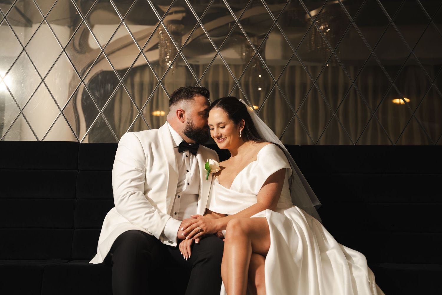 A newlywed couple share an intimate moment sitting together in formal wedding attire during their reception.