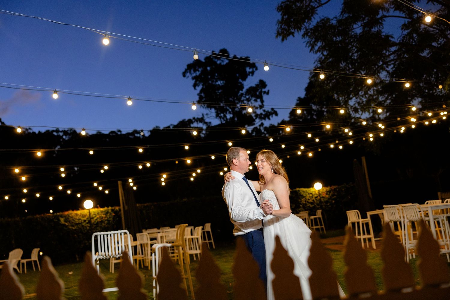 Romantic evening dance at an outdoor wedding reception with string lights illuminating the garden setting.