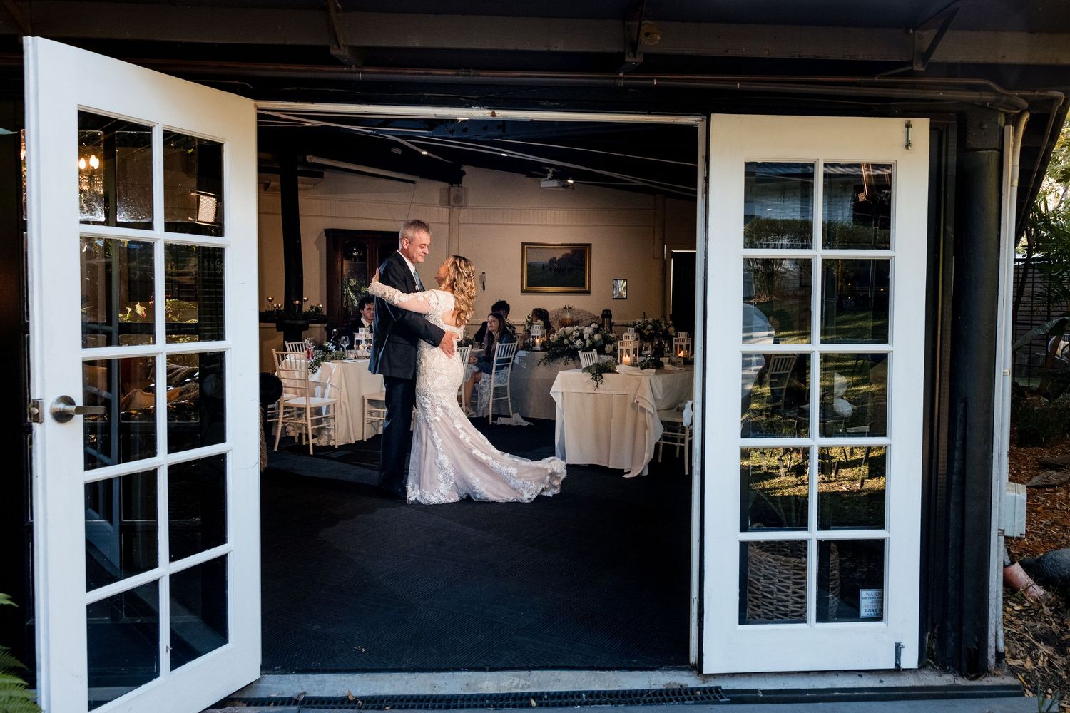 A couple shares their first dance at a wedding reception viewed through elegant French doors opening to a dining room.