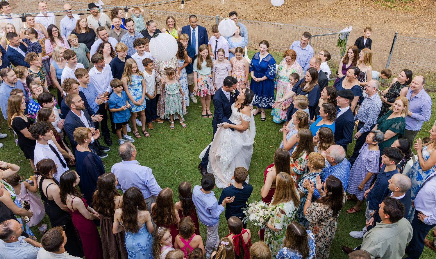 Aerial view of outdoor wedding ceremony guests gathered on a grassy lawn during golden hour.