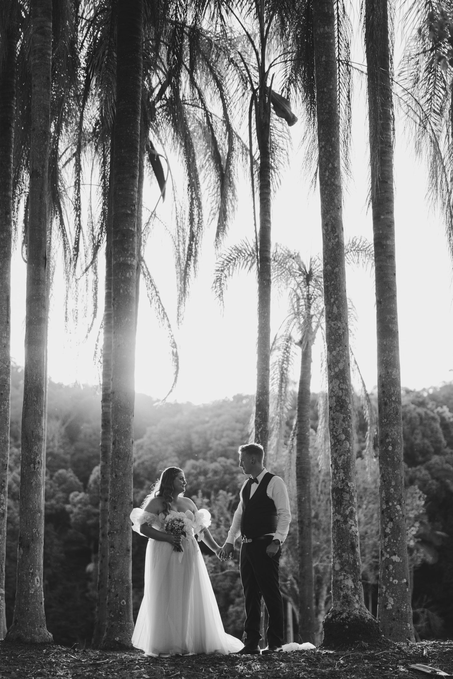 Black and white photo of a bride and groom holding hands among tall palm trees with sunlight streaming through.