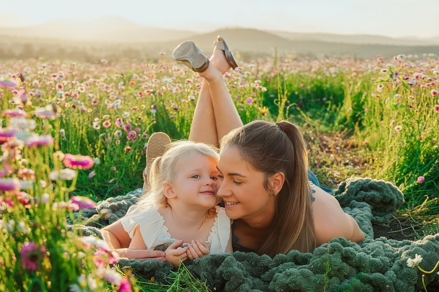Mother and young daughter lying on a green blanket in a blooming wildflower meadow, smiling and touching noses on a sunny day.