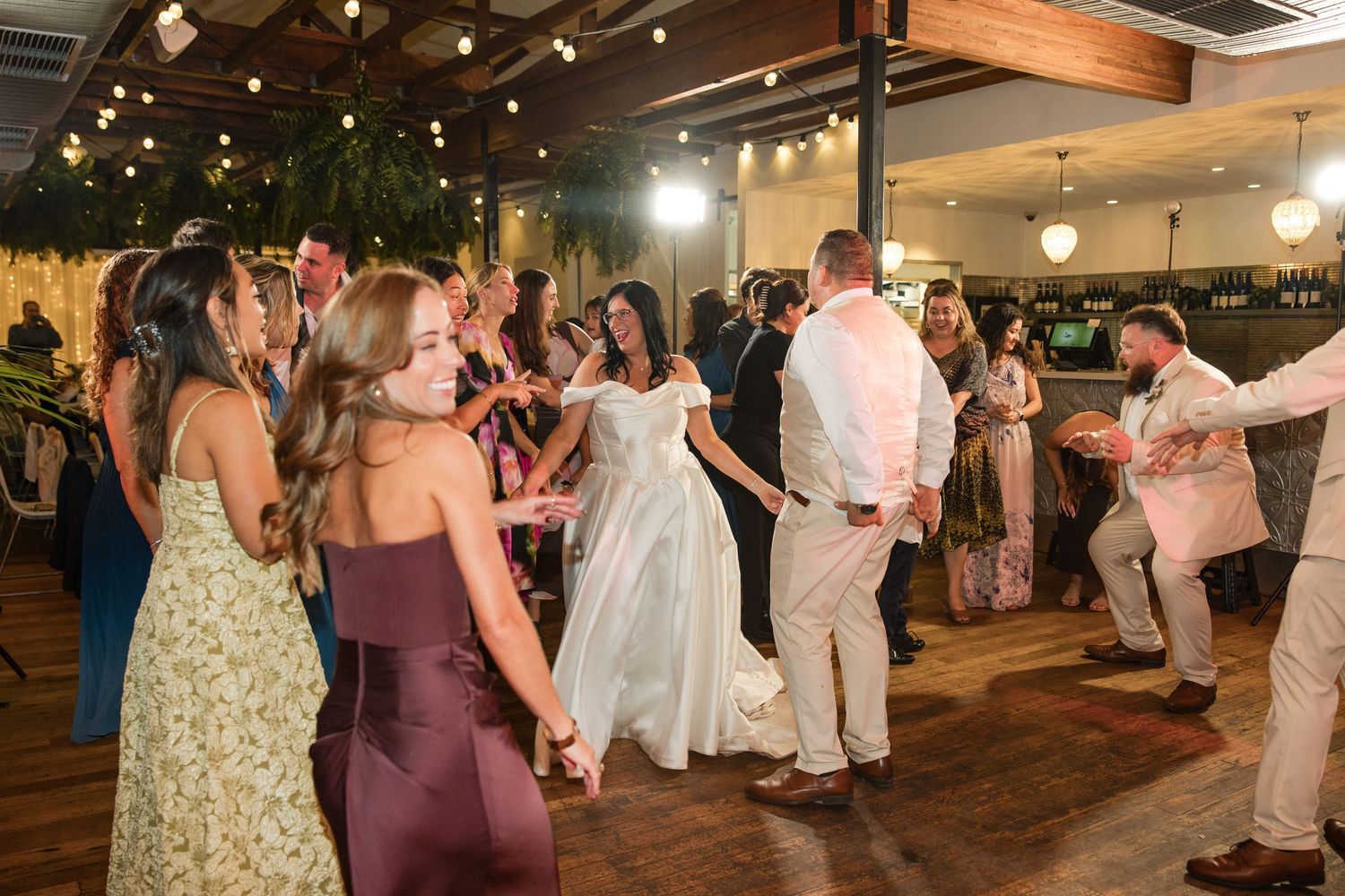Bride and groom dancing joyfully with wedding guests at a elegant indoor reception venue with string lights.