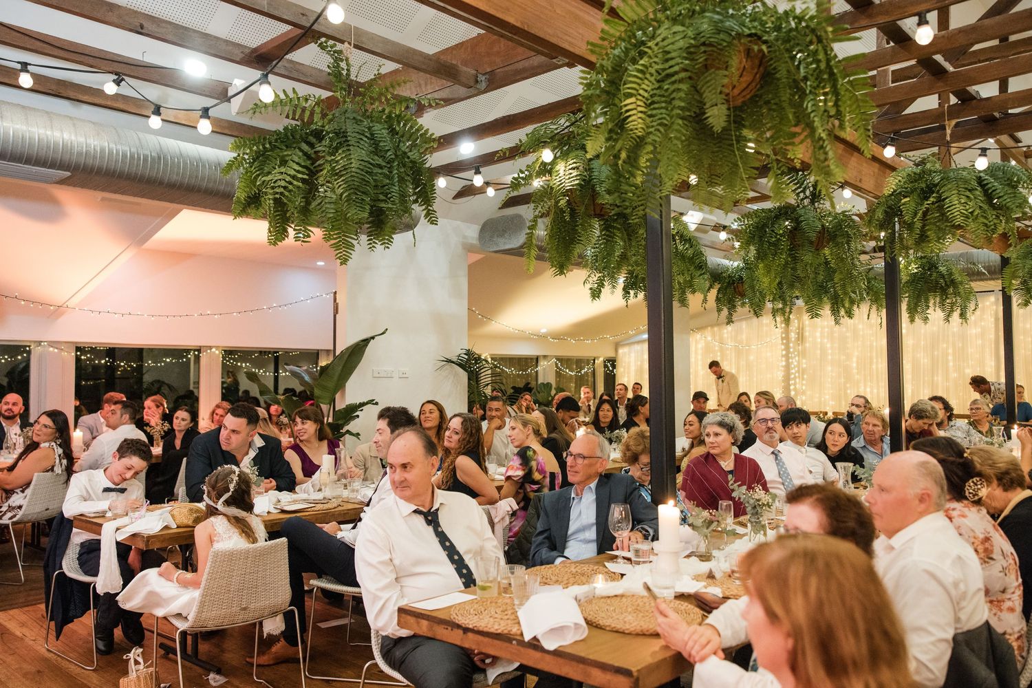 Elegant wedding reception with guests seated at long tables under lush hanging fern arrangements in a warmly lit venue.