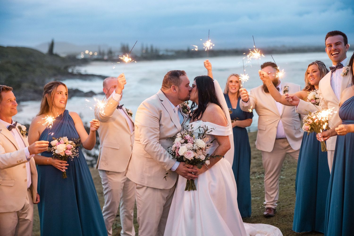 Bride and groom share a kiss surrounded by wedding party holding sparklers near a scenic waterfront at dusk.