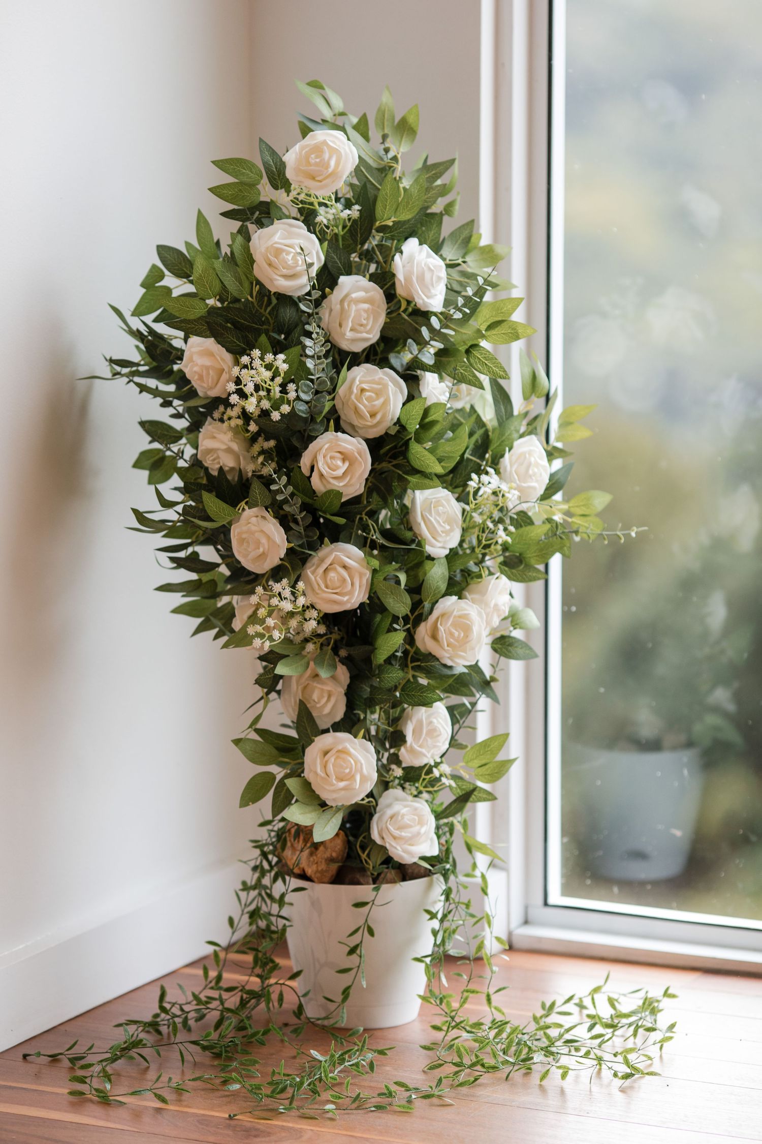 Tall white rose floral arrangement with green foliage in a white pot, placed in a sunlit corner near a window.