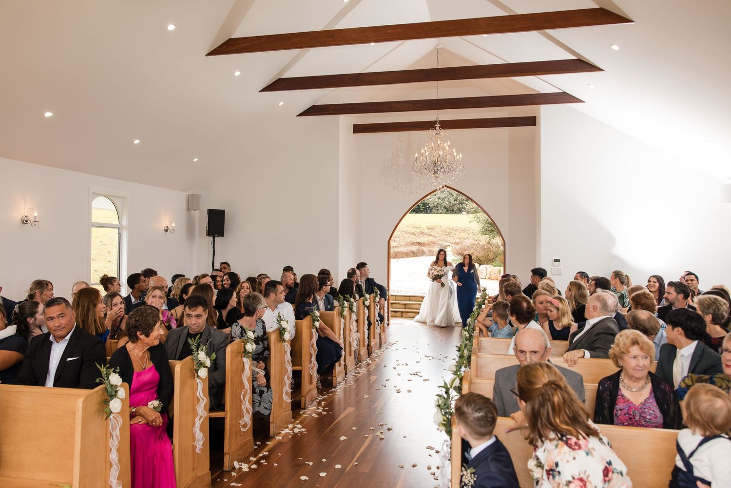 Wedding ceremony in a white chapel with guests seated in wooden pews as couple stands at the altar.