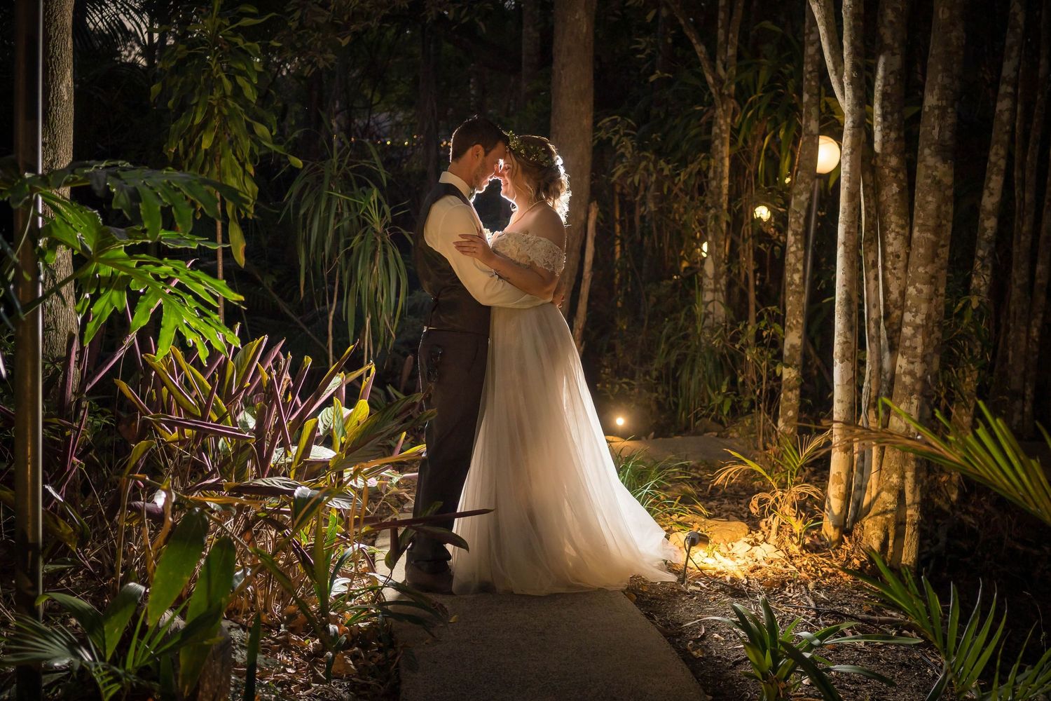 A couple in wedding attire shares a romantic moment in a beautifully lit forest setting at night.