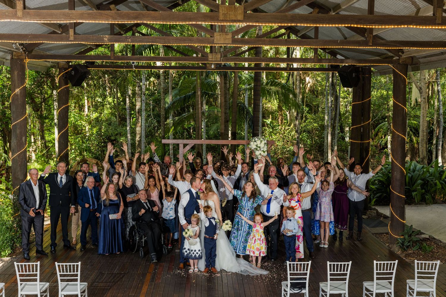 Large group of wedding guests pose together under a covered outdoor venue surrounded by lush tropical forest.
